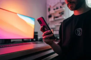 A man holding a smartphone in a bedroom with a colourful monitor behind