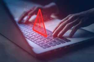 Hands typing on a laptop with a glowing red warning triangle indicating a cybersecurity alert or system vulnerability.