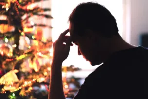 A man sitting in front of a decorated Christmas tree, appearing thoughtful and stressed, highlighting the emotional impact of the festive season