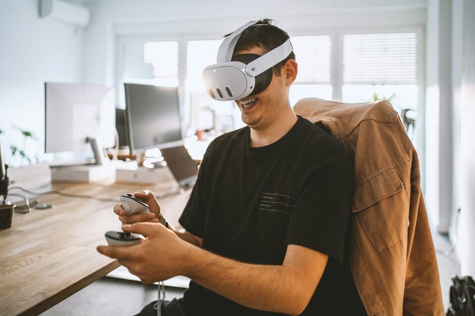 A man using a virtual reality headset sat at a desk with handheld controllers