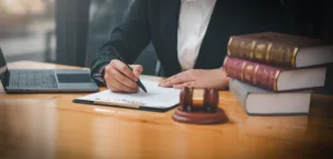 An image of somebody signing a document with a gavel and stack of books in frame.