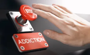 Hand reaching to press a red stop button labeled 'STOP' above a metal plate reading 'ADDICTION' on a black surface